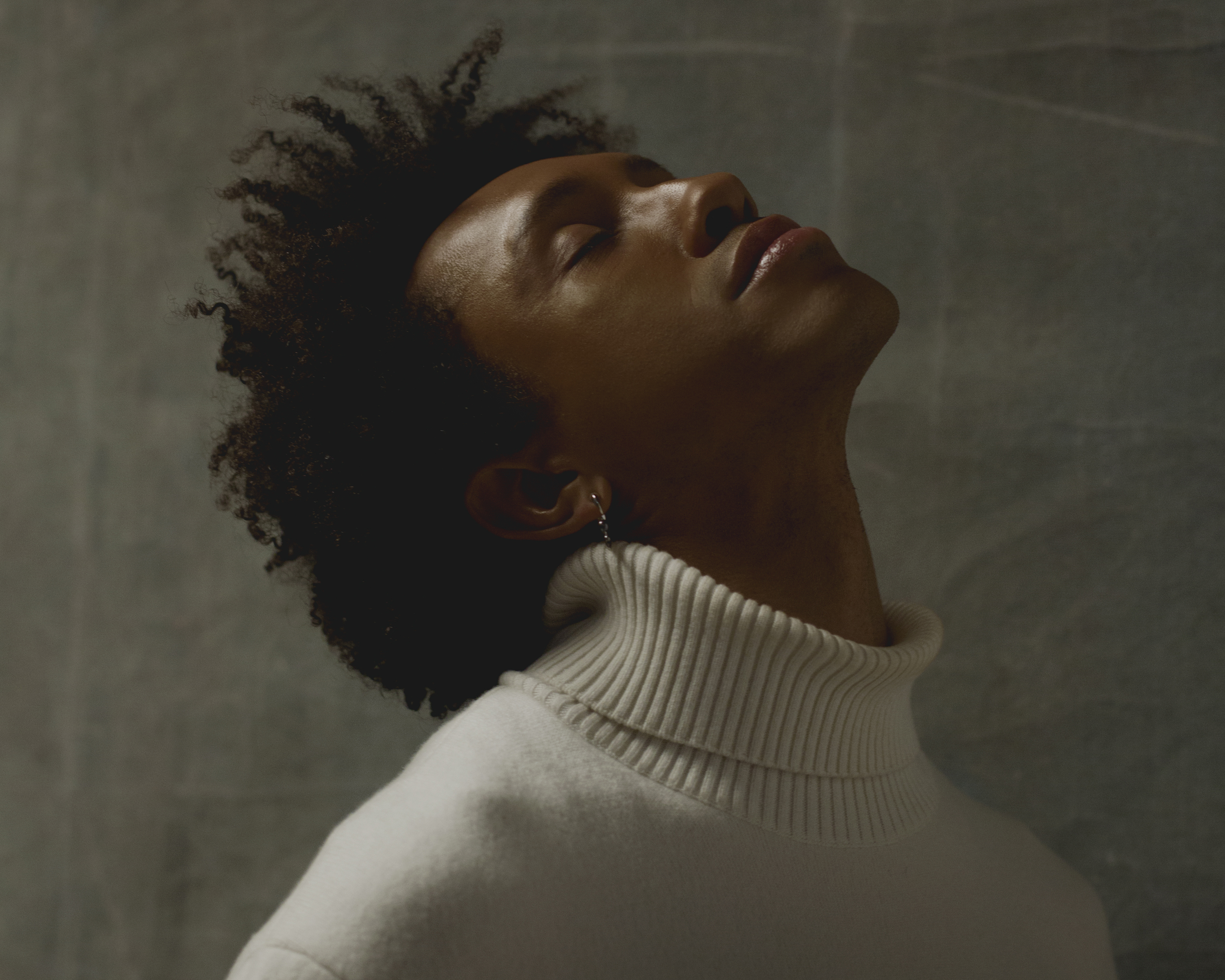 Portrait of Miles Greenberg in a white turtleneck sweater against a dark green wall with his eyes closed and head tilted upwards.