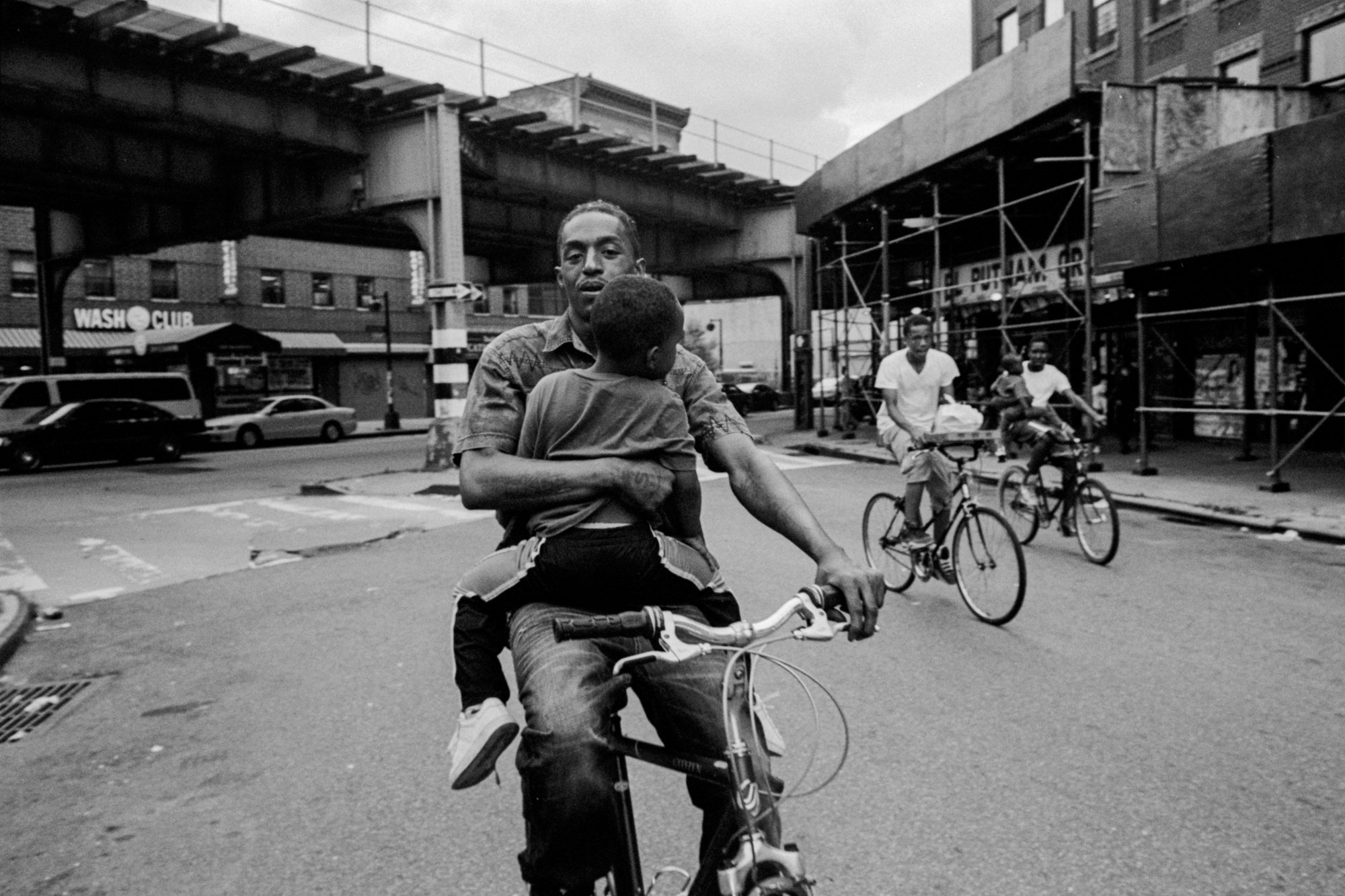Black and white film photograph by Andre. D Wagner of a man holding a child while riding a bike in a city.