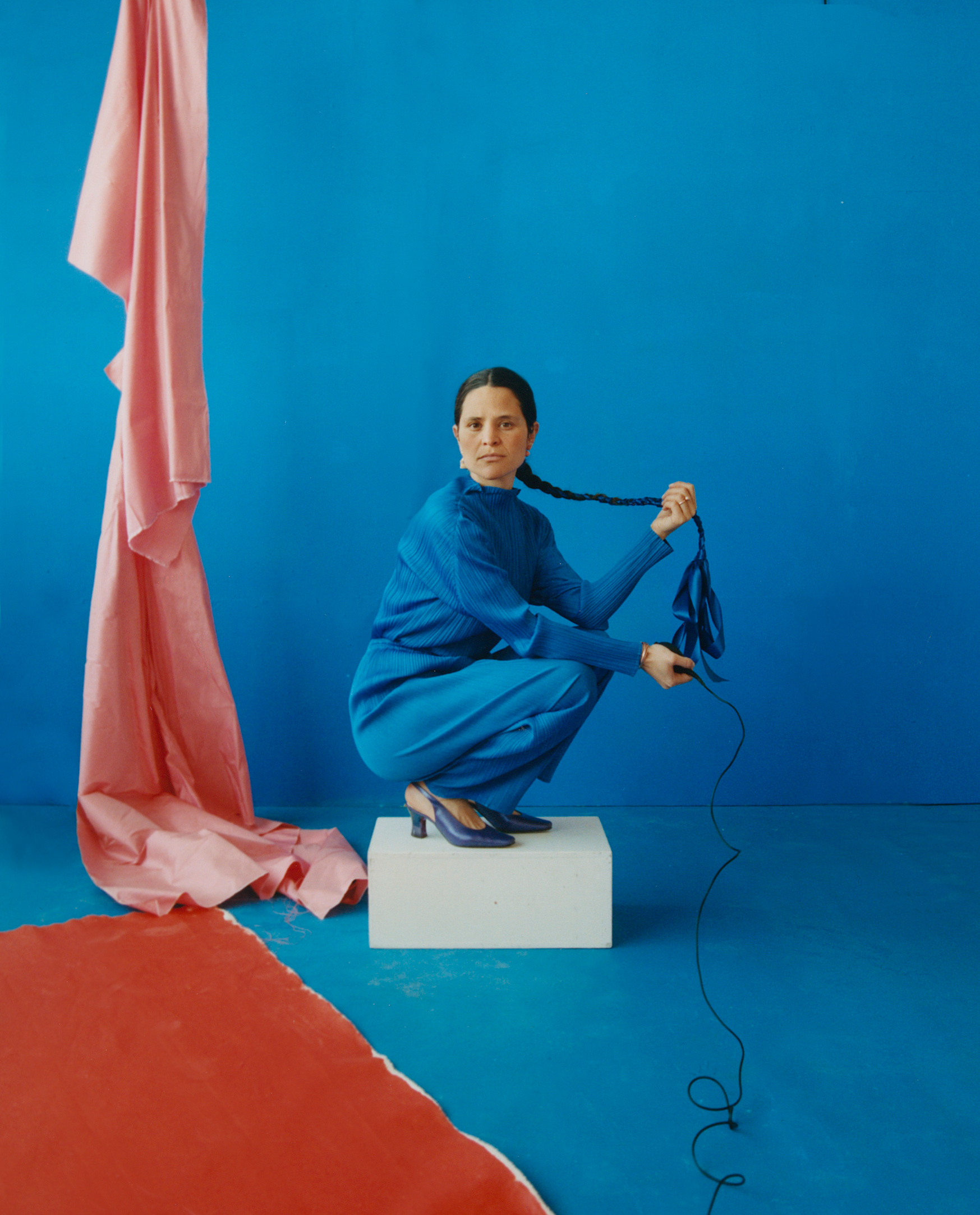 Portrait of Camila Falquez crouching on a white apple box, wearing a long blue gown, holding her braided hair in her hand. The background is blue with a red fabric draped in the left portion of the frame.