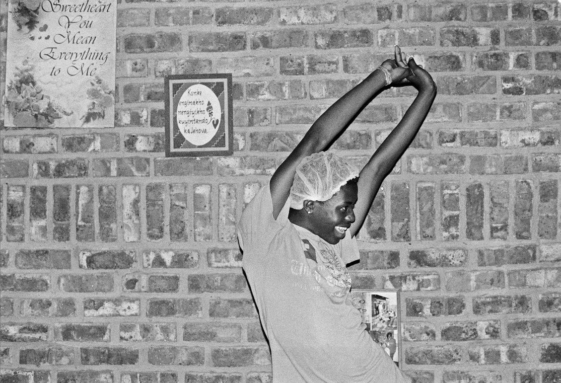 Black and white image of woman standing against brick wall with her arms stretched above her head, smiling to the right.