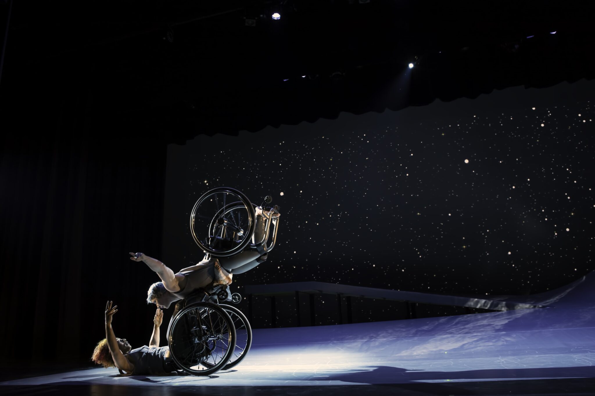 Two dancers in wheelchairs on a blue stage with a starry backdrop. This is Kinetic Light by Alice Sheppard.