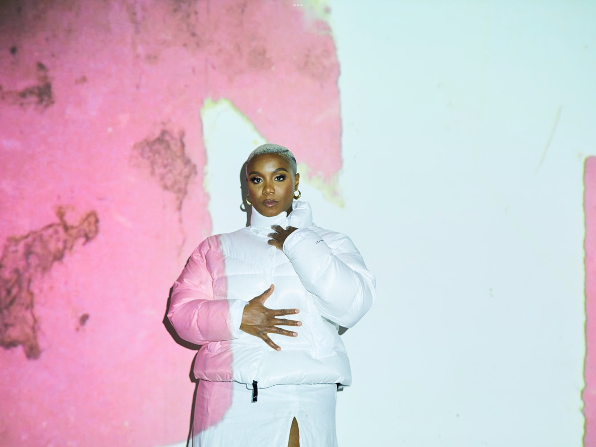 Woman wearing white puffer coat stands against a white wall with pink textures being projected on top of her face.